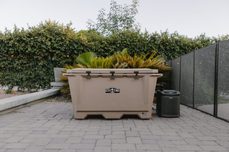 Picture of The Wide Grizzly 450 Cold Plunge Tub sitting on a patio made of brick pavers. The cold plunge lid is closed and latched, and the chiller is attached. There is a hedge behind the tub with some palm-like foliage.